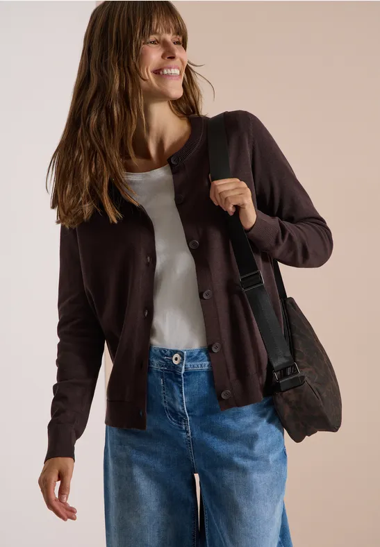 Frau in Jeans, weißem Shirt und brauner Strickjacke mit Schultertasche, lächelt locker vor neutralem Hintergrund.