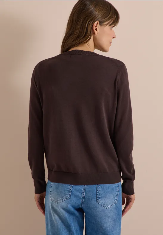 Frau von hinten in braunem Pullover und blauen Jeans vor neutralem Hintergrund.