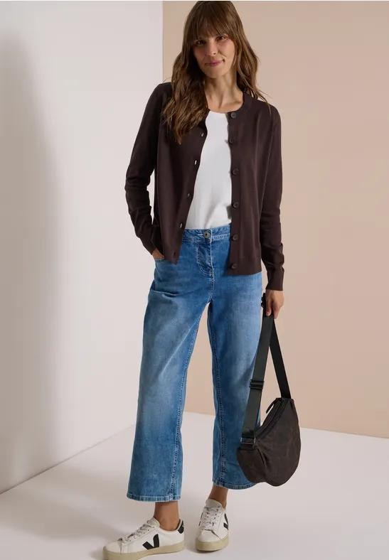 Frau mit braunen Haaren trägt weißes Shirt, braune Jacke, weite Jeans, weiße Sneaker und eine schwarze Tasche.