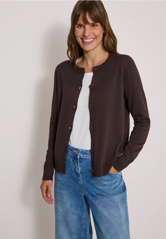 Eine lächelnde Frau trägt eine braune Strickjacke, weißes Shirt und blaue Jeans.