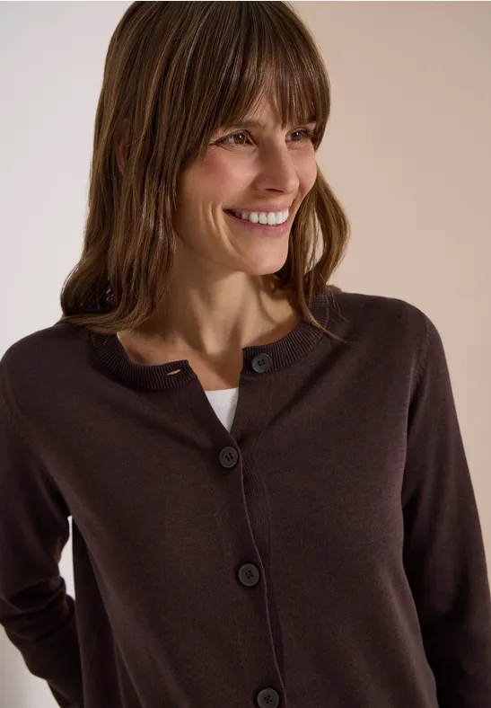 Eine Frau trägt eine dunkelbraune Strickjacke, hat braune Haare und lächelt vor hellem Hintergrund.