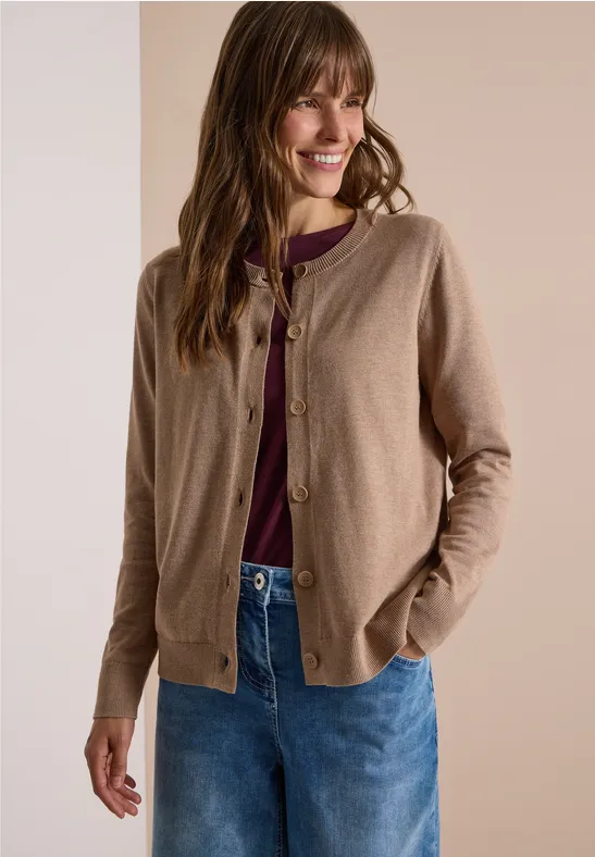 Frau trägt beige Strickjacke, weinrotes Shirt und blaue Jeans, lächelnd mit Händen in den Taschen.