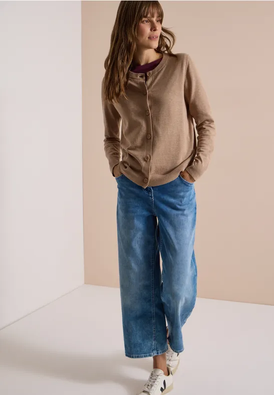 Frau lehnt lässig an Wand, trägt beigen Hoodie, weite Jeans und helle Sneakers.