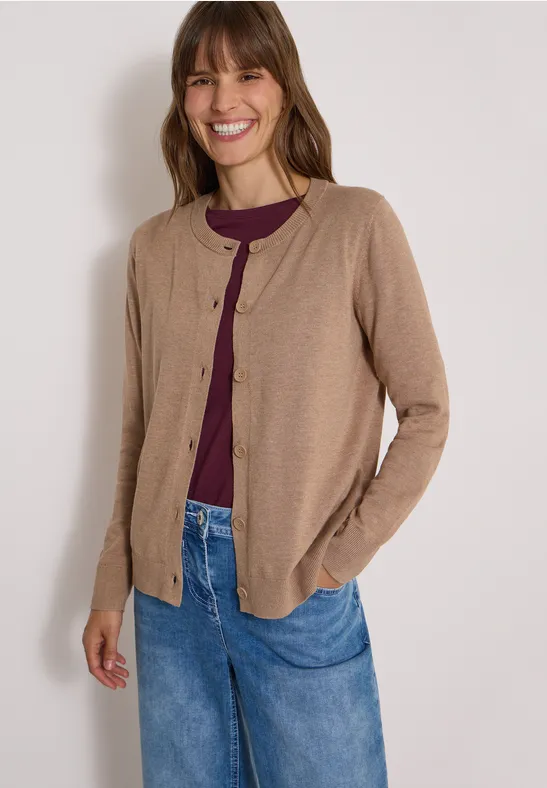 Eine lächelnde Frau trägt eine beige Strickjacke über einem lila Oberteil und Jeans.