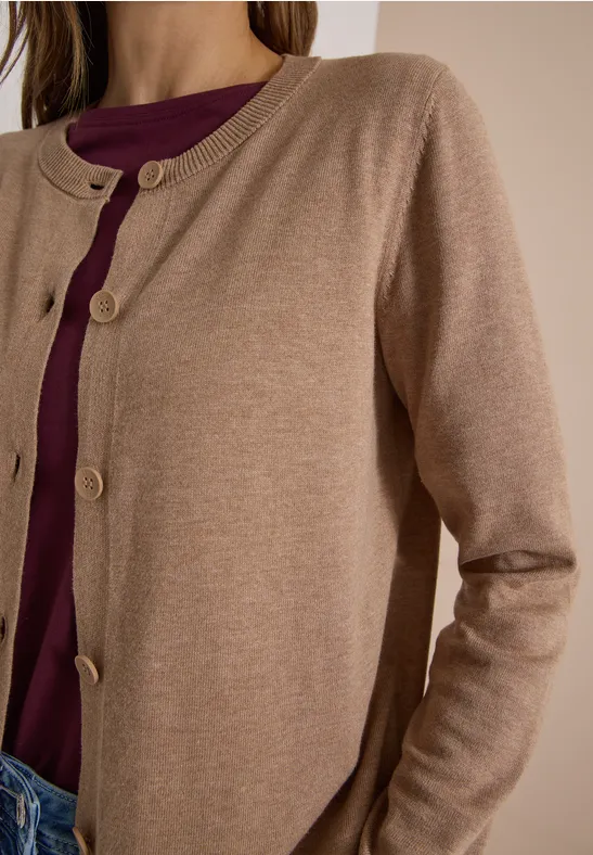 Frau trägt eine beige Strickjacke mit Knöpfen über einem dunkellila Oberteil und blauen Jeans.