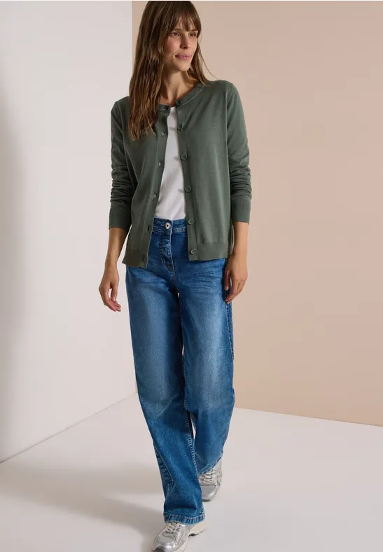 Eine Frau in grüner Strickjacke, weißem Shirt, blauen Jeans und Turnschuhen.