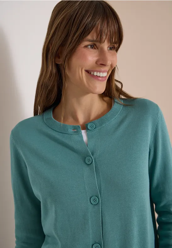 Frau in einem grüntürkisen Strickcardigan lächelt und blickt nach rechts.