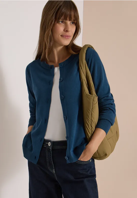 Eine junge Frau trägt eine blaue Strickjacke, weiße Bluse, Jeans und eine grüne Tasche.