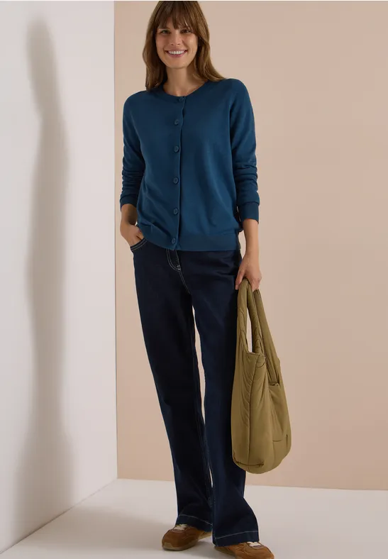 Frau in blauer Strickjacke und dunklen Jeans trägt eine grüne Tasche und lächelt vor schlichtem Hintergrund.