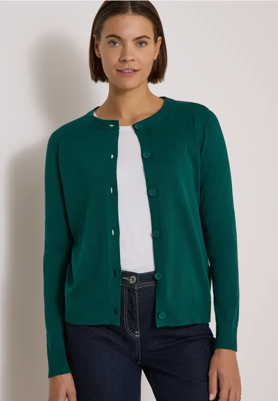 Frau trägt grünen Cardigan, weißes Shirt und Jeans vor hellem Hintergrund.