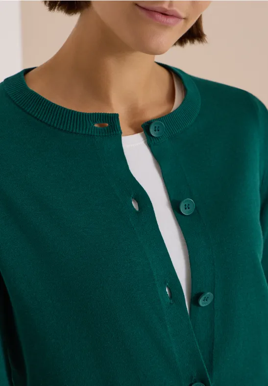 Grüne Damen-Strickjacke mit Rundhalsausschnitt und Knopfleiste, getragen über weißem Oberteil.