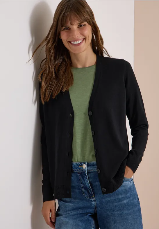 Eine Frau steht lächelnd in Jeans, grünem T-Shirt und schwarzer Strickjacke vor einer Wand.