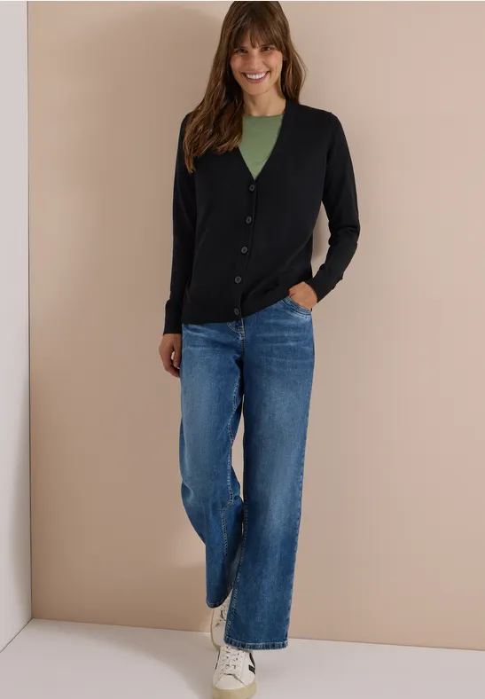Eine lächelnde Frau trägt schwarzen Cardigan, grünes Top, weite blaue Jeans und weiße Sneaker vor beiger Wand.