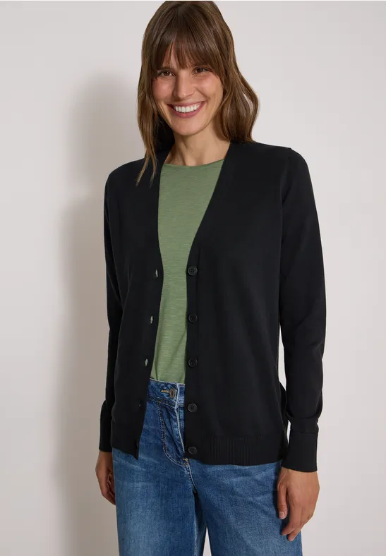 Eine Frau mit schwarzer Strickjacke, grünem Oberteil und blauen Jeans lächelt in die Kamera.