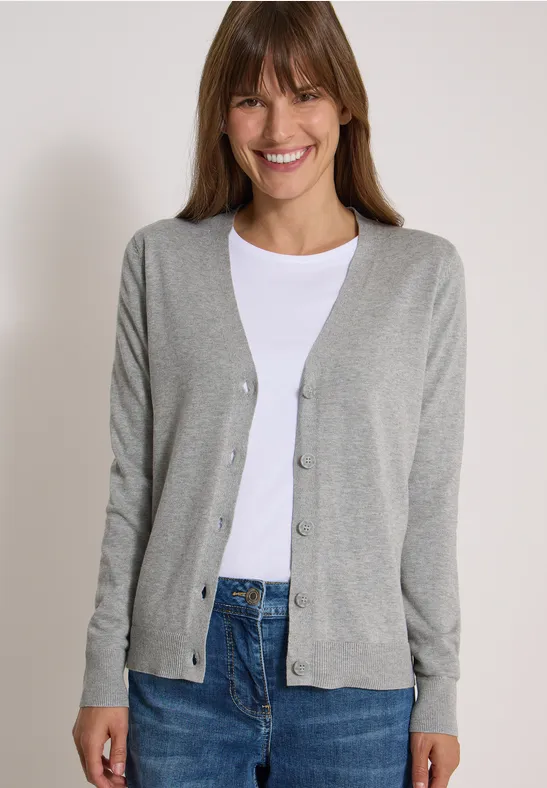 Frau mit hellem Cardigan, weißem Shirt und Jeans lächelt vor hellem Hintergrund.