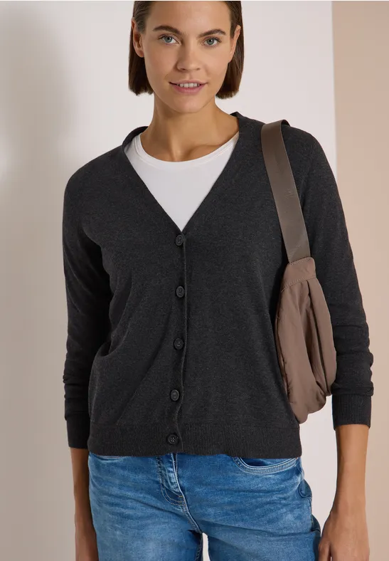 Frau trägt dunkelgrauen Cardigan, weißes Shirt, blaue Jeans und Umhängetasche vor neutralem Hintergrund.