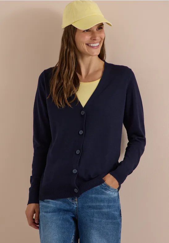 Frau in dunkelblauer Strickjacke, gelber Cap und Jeans lächelt.