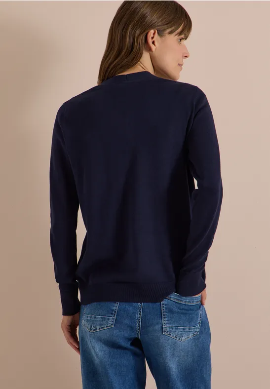 Frau von hinten in dunkelblauem Pullover und Jeans vor beigem Hintergrund.