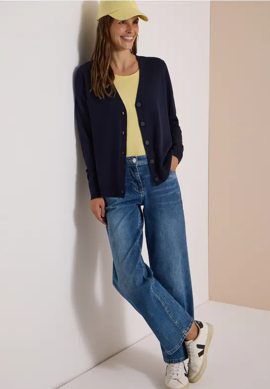 Junge Frau lehnt an Wand, trägt gelbes Top, dunkelblauen Cardigan, Jeans, Cap und weiße Sneaker.