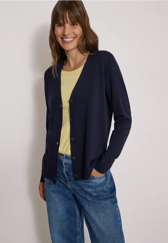 Frau trägt dunkelblauen Cardigan, gelbes T-Shirt und blaue Jeans, schaut lächelnd in die Kamera.