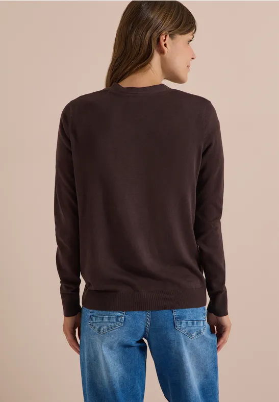 Frau von hinten in dunkelbraunem Pullover und blauen Jeans vor hellem Hintergrund.
