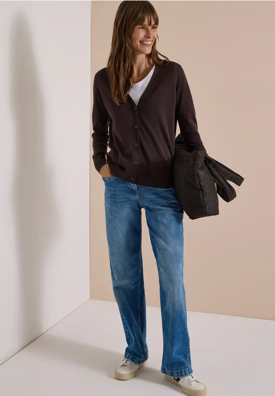 Frau in Cardigan, Jeans und Sneakern hält eine Jacke vor neutralem Hintergrund.