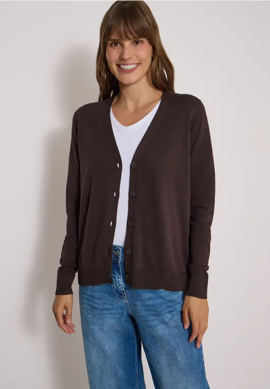 Frau in brauner Strickjacke, weißem T-Shirt und Jeans, lächelnd vor neutralem Hintergrund.