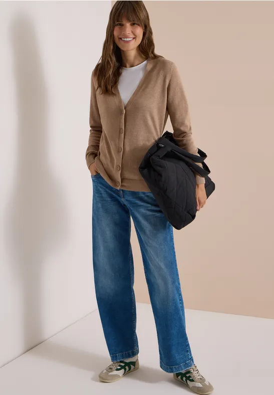 Frau in beiger Strickjacke, weißem T-Shirt, weitem Jeans und Sneakers trägt eine dunkle Jacke vor neutralem Hintergrund.