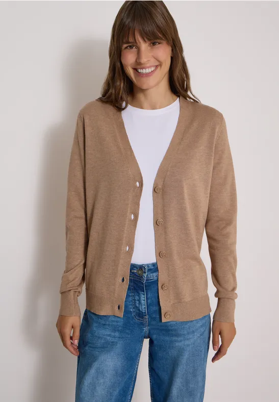 Frau im hellbraunen Cardigan über weißem T-Shirt und blauen Jeans lächelt in die Kamera.