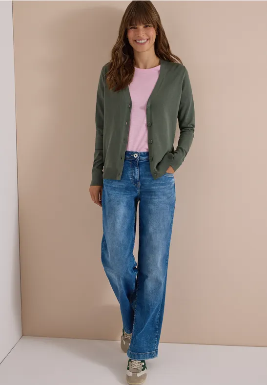 Vrouw in groen vest, roze t-shirt en wijde jeans poseert voor een beige muur.