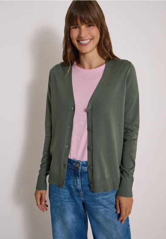 Een vrouw draagt een groene cardigan over een roze shirt en een blauwe spijkerbroek.