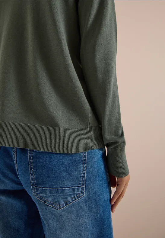 Achteraanzicht van een persoon in een groen shirt met lange mouwen en een blauwe spijkerbroek tegen neutrale achtergrond.
