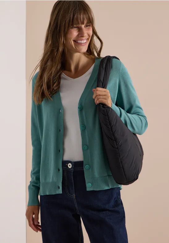 Frau in türkiser Strickjacke, weißem Shirt und dunklen Jeans mit schwarzer Schultertasche lächelnd.