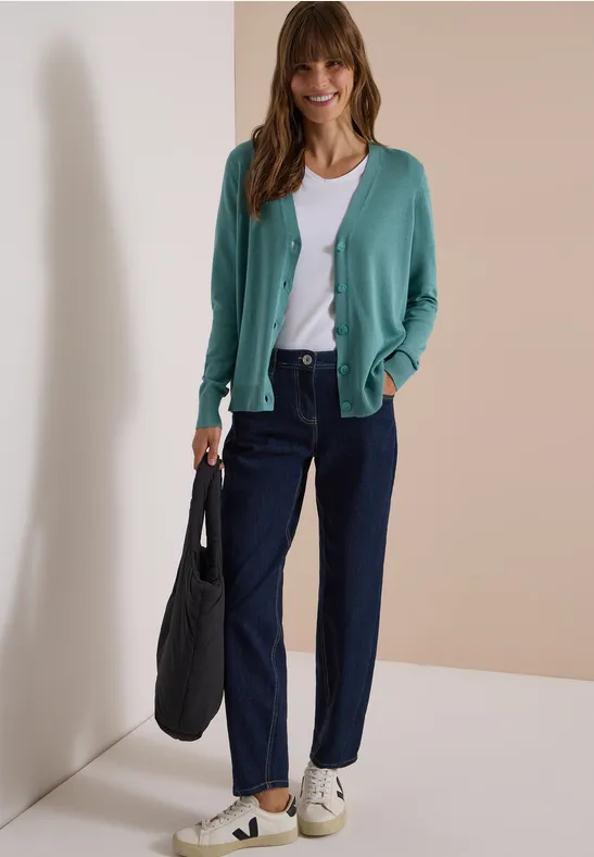 Eine Frau in grünem Cardigan, weißem Top, dunklen Hosen und Sneakern hält eine schwarze Tasche vor beigem Hintergrund.