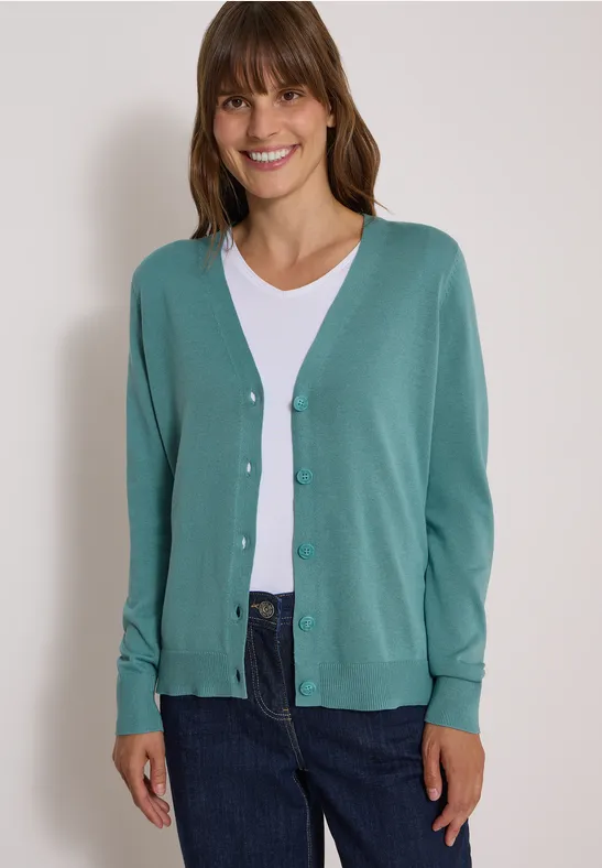 Frau mit grüner Strickjacke, weißem Shirt und dunkler Jeans, lächelnd vor neutralem Hintergrund.