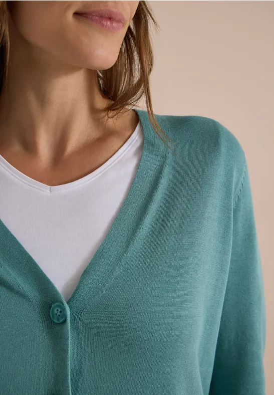 Eine Frau trägt eine türkisfarbene Strickjacke über einem weißen Top.
