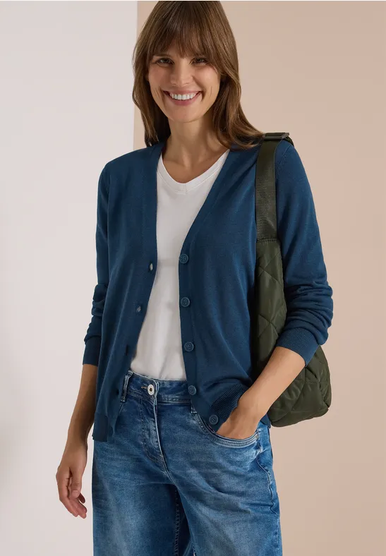 Frau in weißem T-Shirt, blauem Cardigan und Jeans trägt grüne Tasche und lächelt.
