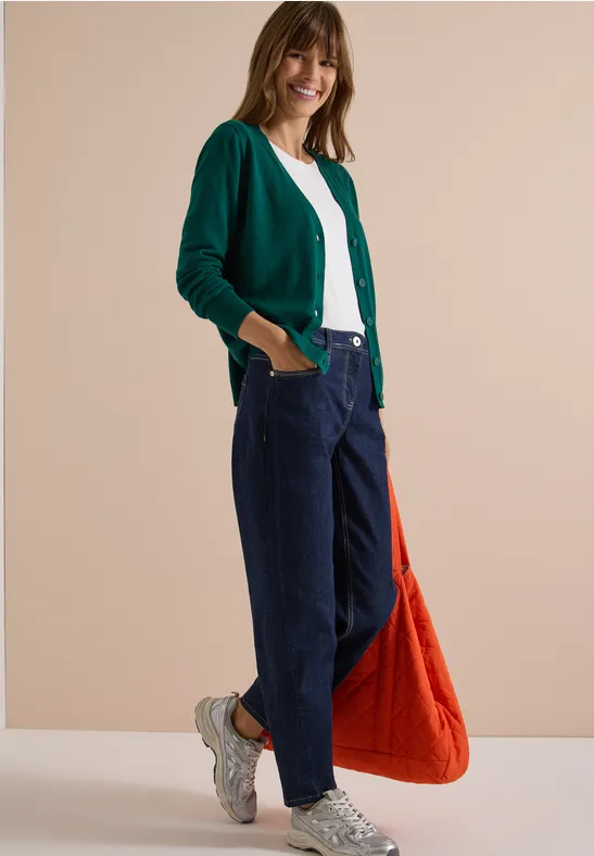 Frau in grünem Cardigan, weißem Shirt und blauen Jeans mit weißen Sneakern und orangener Tasche vor neutraler Wand.