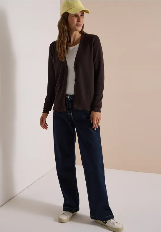 Junge Frau in weiter Jeans, weißem T-Shirt, dunklem Cardigan und gelber Cap lehnt an einer Wand.