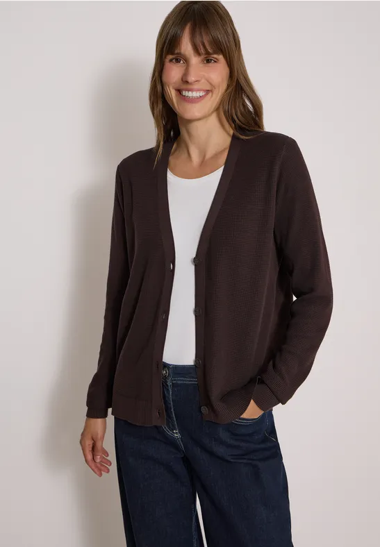 Eine lächelnde Frau trägt eine dunkelbraune Strickjacke über weißem T-Shirt und blauen Jeans.