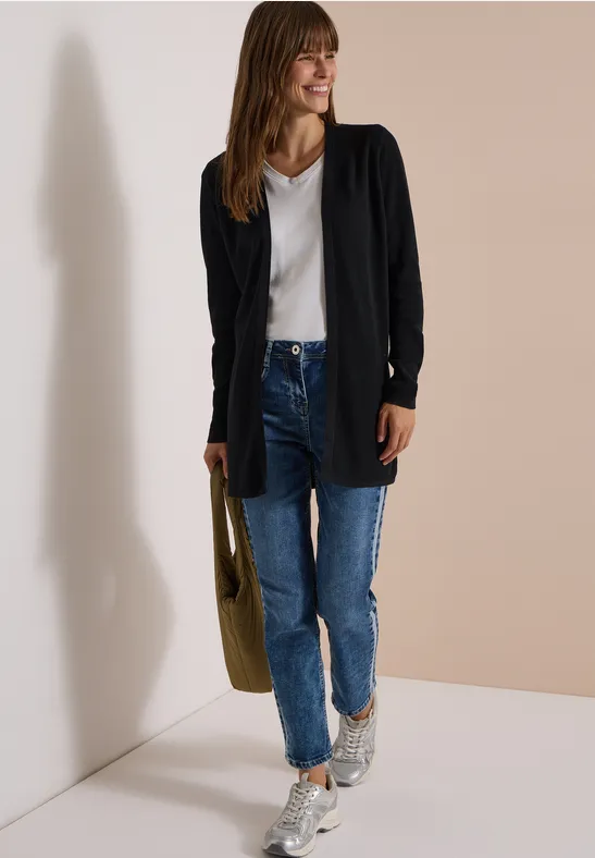 Frau trägt schwarzen Cardigan, weißes V-Neck-Shirt, blaue Jeans, Sneakers und hält olivgrüne Tasche.