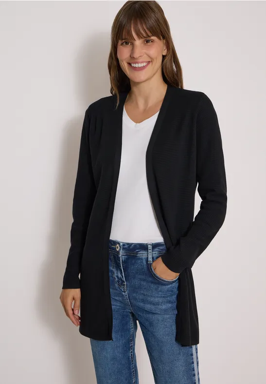 Eine lächelnde Frau trägt eine schwarze Strickjacke, weiße Bluse und blaue Jeans.