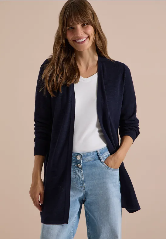 Eine lächelnde Frau mit langen Haaren trägt ein weißes Top, blaue Jeans und einen dunkelblauen Cardigan vor beigem Hintergrund.