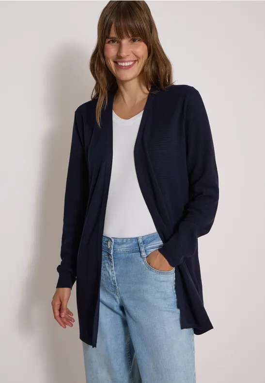 Frau mit braunen Haaren, lächelnd in weißem T-Shirt, blauer Strickjacke und Jeans vor hellem Hintergrund.