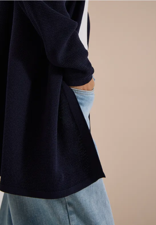 Ein dunkelblauer Cardigan über weißem Top, kombiniert mit hellblauen Jeans und Hand in Hosentasche.
