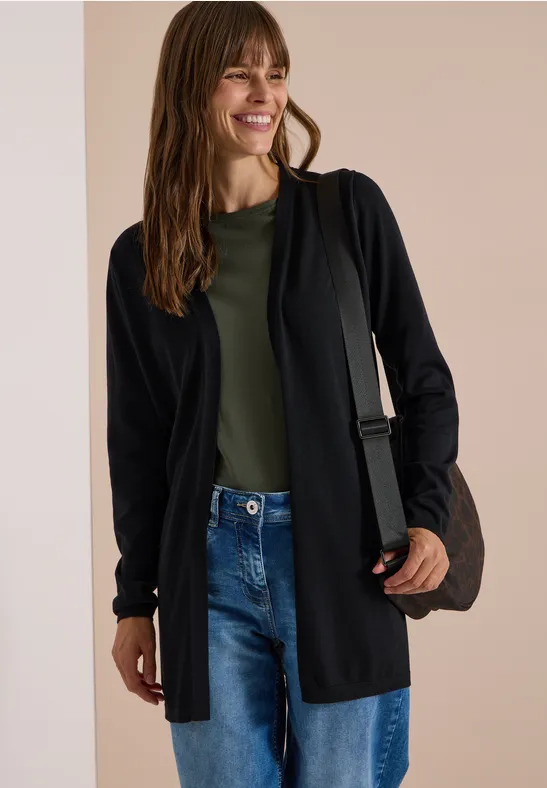 Eine Frau trägt einen schwarzen Cardigan, grünes Shirt, Jeans und lächelt, mit Tasche über der Schulter.