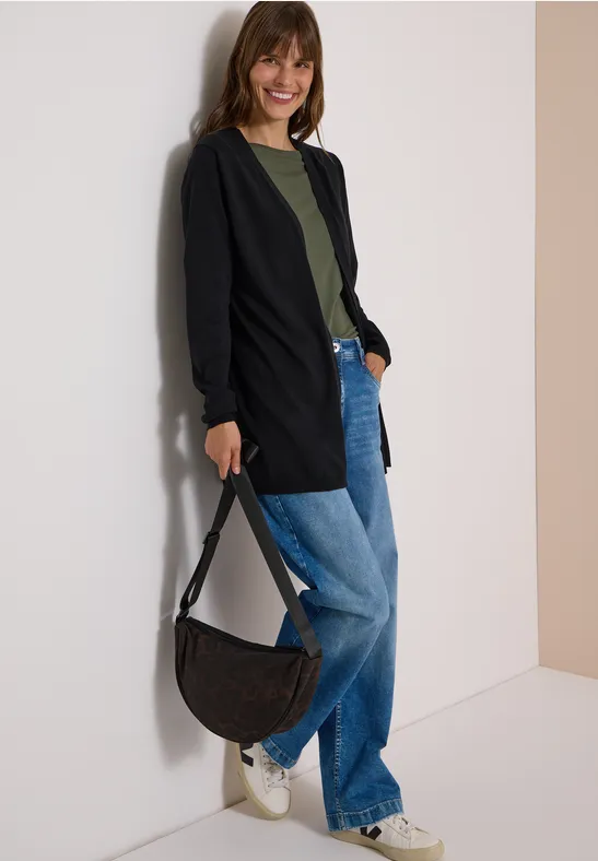 Frau lehnt an der Wand, trägt schwarzen Cardigan, grünes Shirt, weite Jeans, weiße Sneaker und schwarze Tasche.