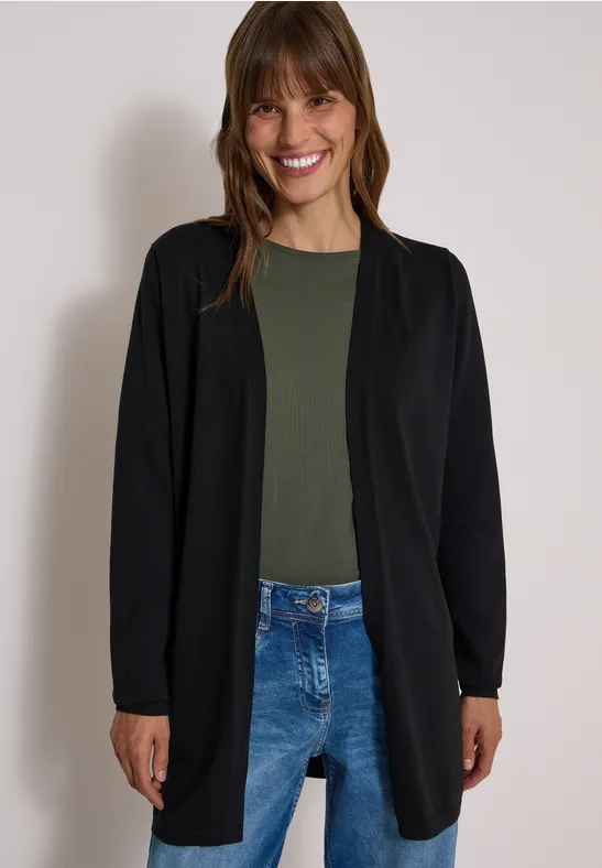 Eine lächelnde Frau in schwarzer Strickjacke, olivgrünem Shirt und blauen Jeans vor neutralem Hintergrund.