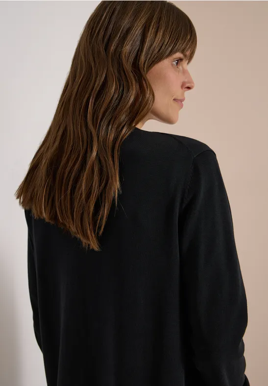 Frau mit langen, welligen braunen Haaren, trägt schwarzen Pullover, steht vor hellem Hintergrund und blickt seitlich.