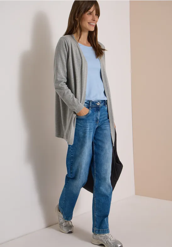 Frau trägt graue Strickjacke, hellblaues Top, weite Jeans und Sneaker, hält eine schwarze Tasche.